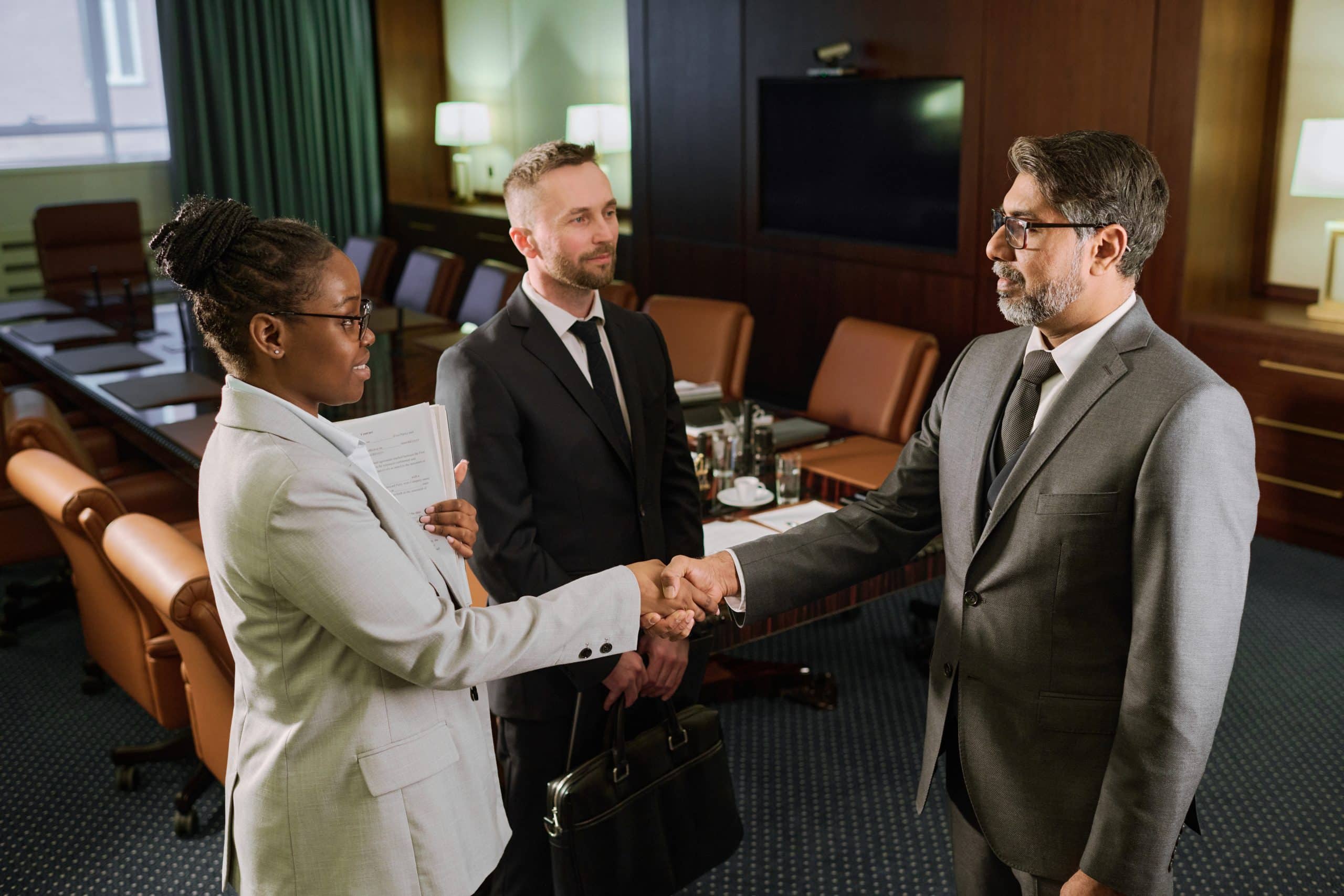 Confident mature businessman in suit looking at young female colleague during handshake while both standing next to male lawyer