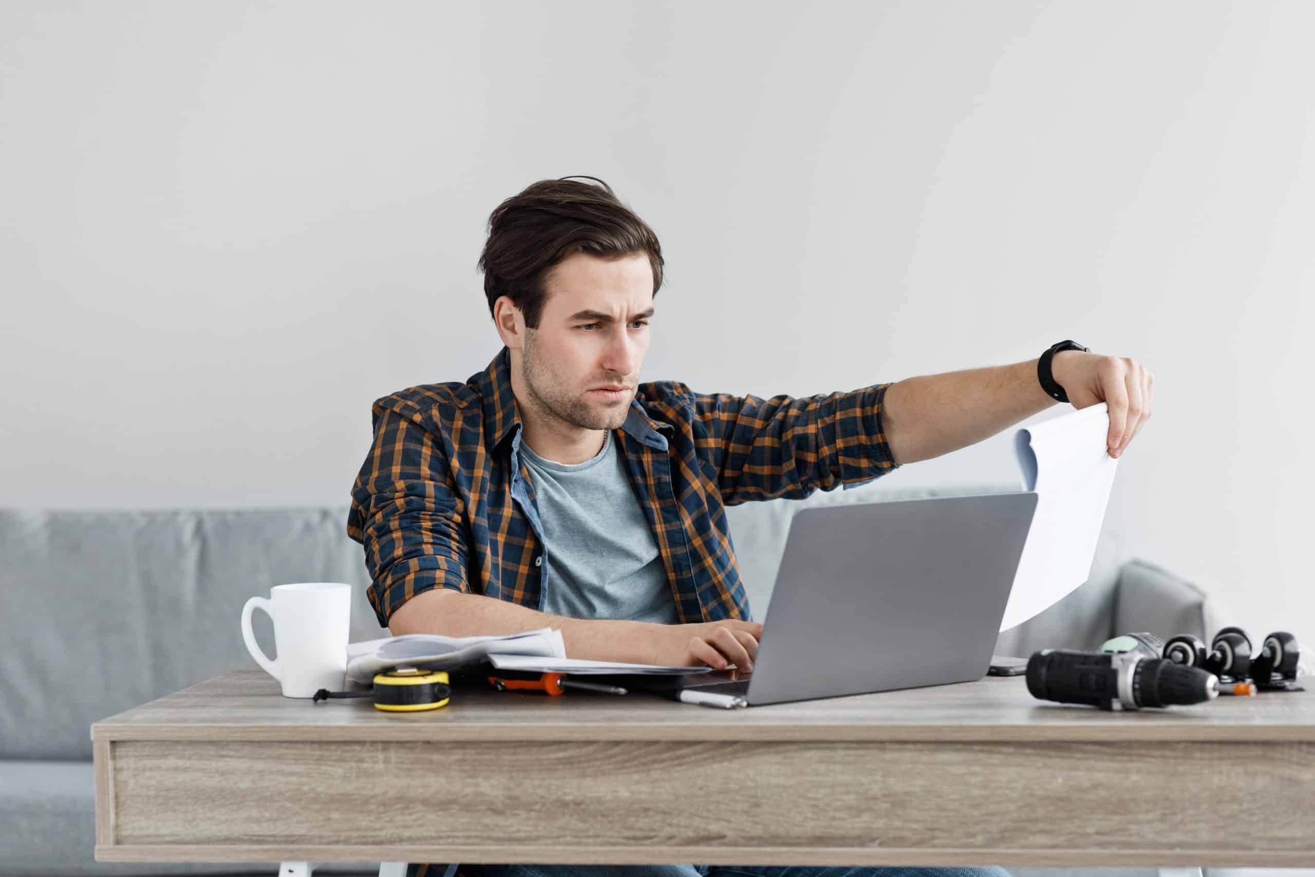 Man reading documents at laptop, home office setup.